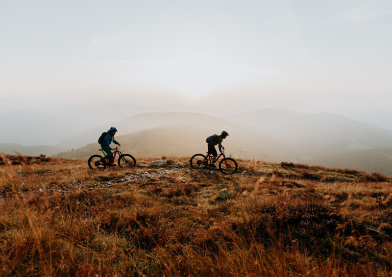 Zwei Mountainbiker fahren im Abendlicht über einen alpinen Trail, mit nebligen Bergkuppen im Hintergrund.