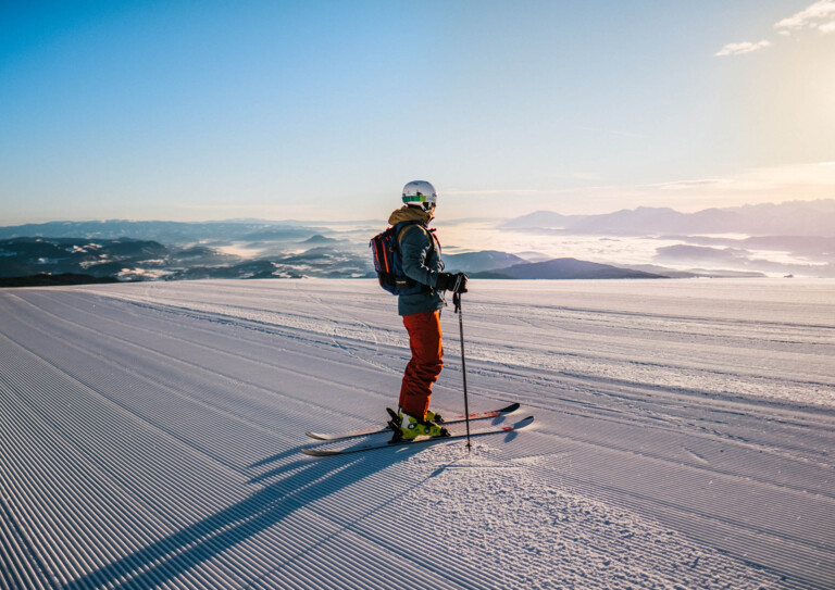 Ein Skifahrer steht auf einer frisch präparierten Piste mit Blick auf eine verschneite Berglandschaft bei Sonnenaufgang.