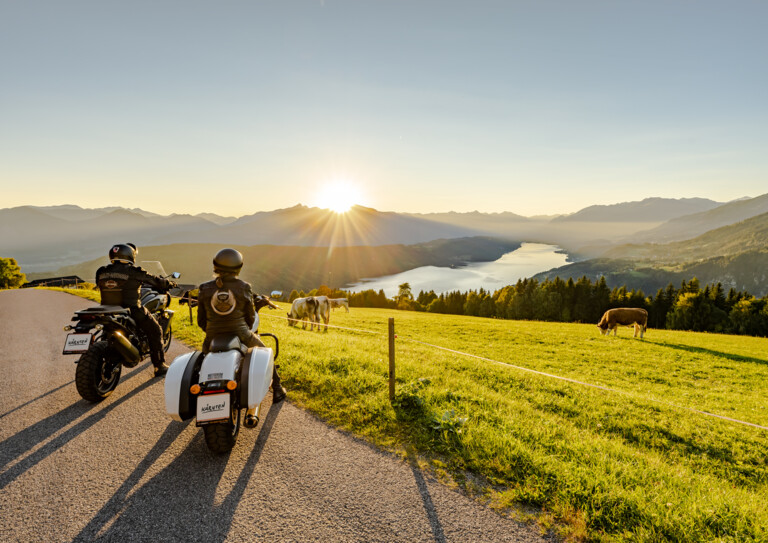 Zwei Motorradfahrer genießen den Sonnenuntergang auf einer malerischen Straße mit Blick auf einen See und Berge. Kühe grasen auf der Wiese.
