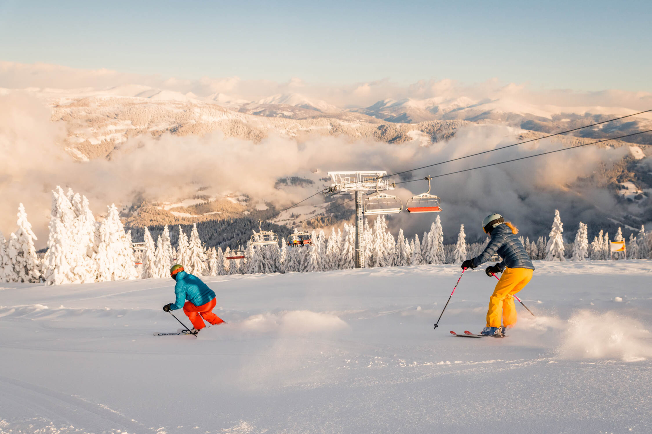 Zwei Personen beim Skifahren auf einer Skipiste der Gerlitzen Alpe, umgeben von verschneiten Bäumen und Bergen im Hintergrund.