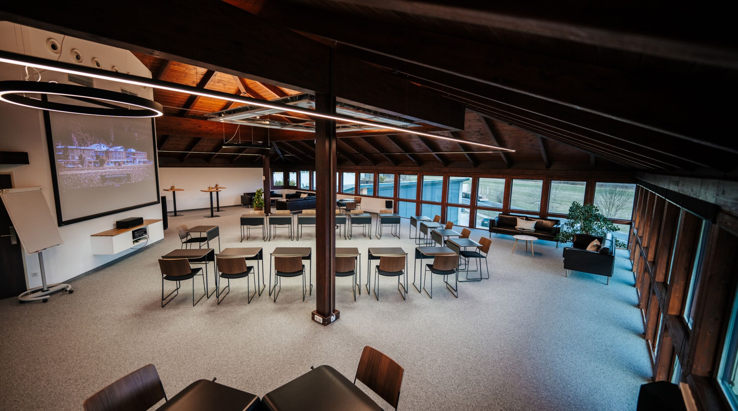 Moderner Seminarraum mit Holzdach, Projektor und Sitzgelegenheiten, ideal für Veranstaltungen. Große Fenster bieten viel Tageslicht und Aussicht.