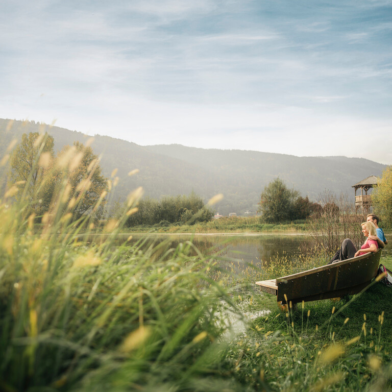 A couple relaxes on a bench on the Slow Trail in the Bleistätter Moor in Villach, surrounded by lush greenery and distant mountains under a clear sky.