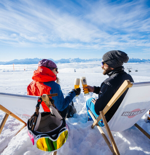 Two people in winter clothing clinking glasses on a snowy mountain, sitting on deck chairs with a scenic view of the Alps.