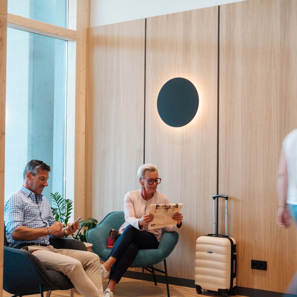 Two people sitting in a modern waiting area, one reading a newspaper. A suitcase is nearby. Two people walk past.