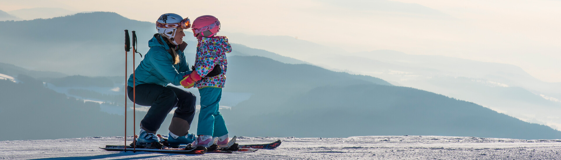 Frau in der Hocke umarmt kleines Mädchen, das auf Skiern steht.