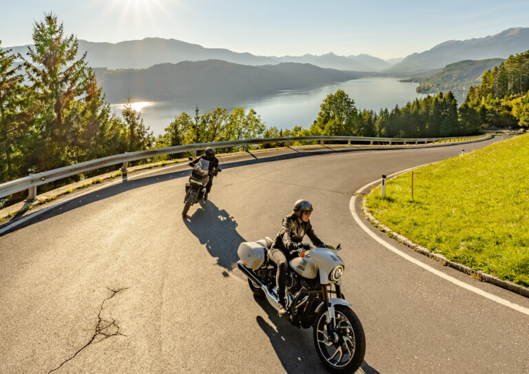 Zwei Motorradfahrer fahren auf einer kurvigen Straße mit Blick auf einen See und Berge im Sonnenschein.