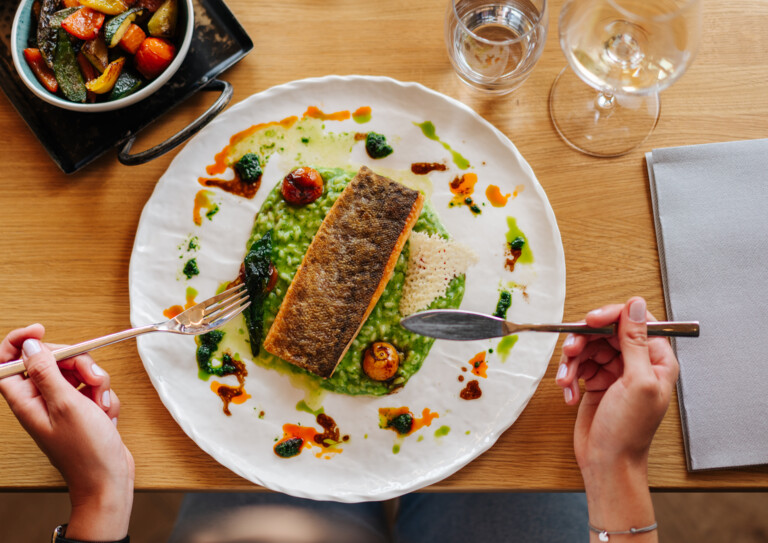 Person genießt gegrillten Fisch auf Risotto, begleitet von gebratenem Gemüse, an einem Tisch mit einem Glas Wein.