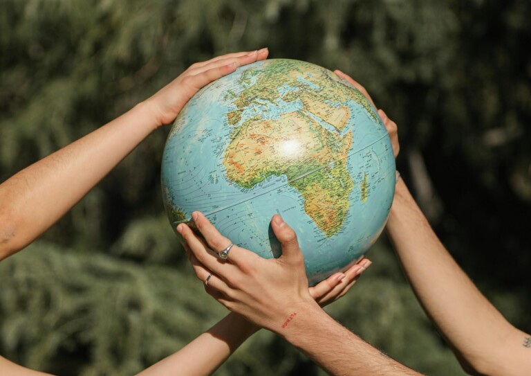 Mehrere Hände halten gemeinsam einen Globus, im Hintergrund befinden sich Bäume.