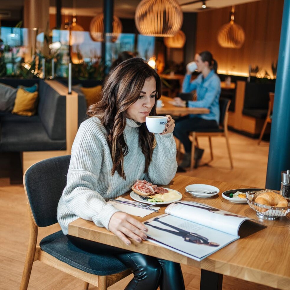 A woman reads a magazine while enjoying coffee and a sandwich in the breakfast area of Hotel Educare.
