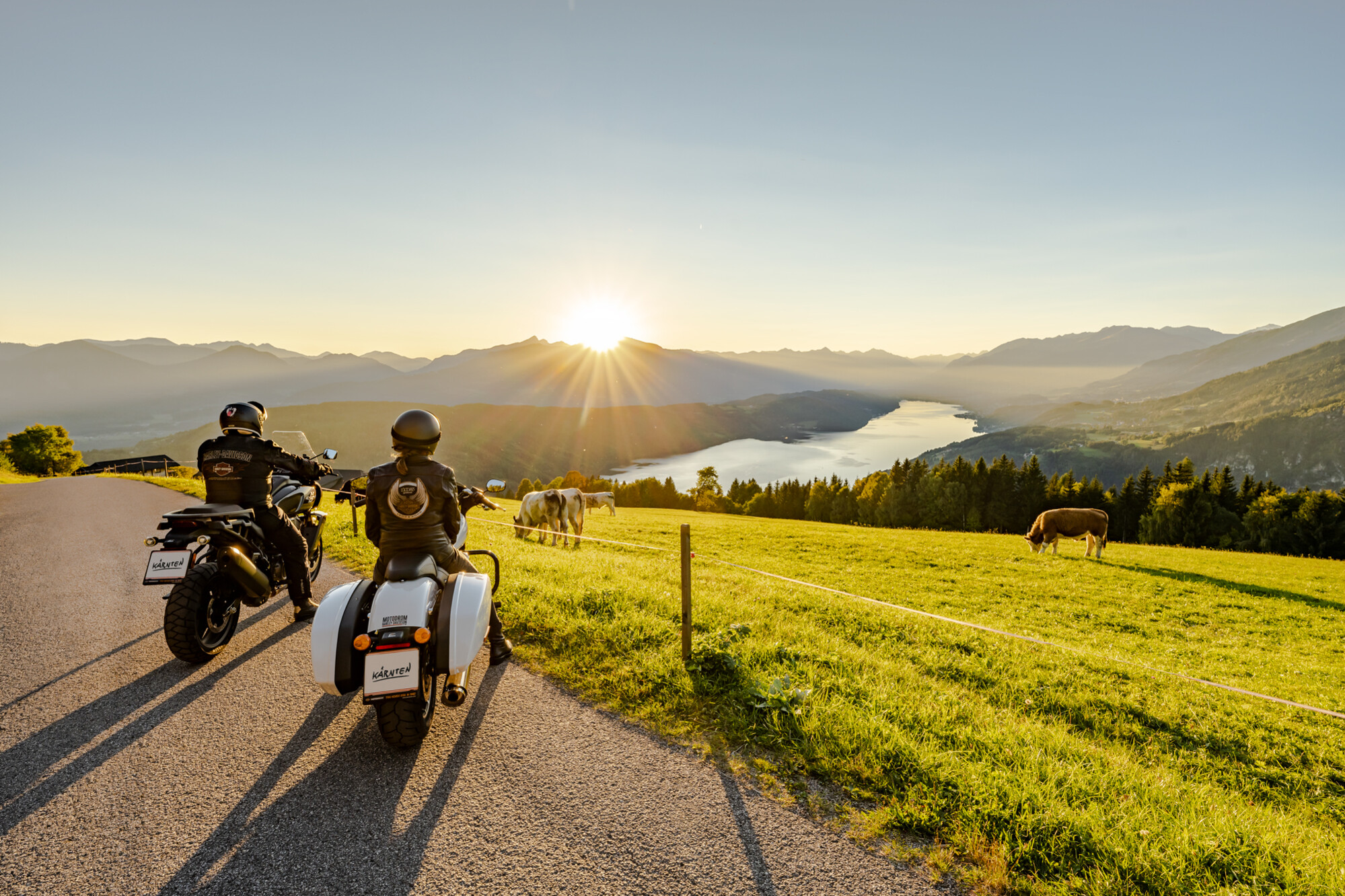 Zwei Motorradfahrer genießen den Sonnenuntergang auf einer malerischen Straße mit Blick auf einen See und Berge. Kühe grasen auf der Wiese.
