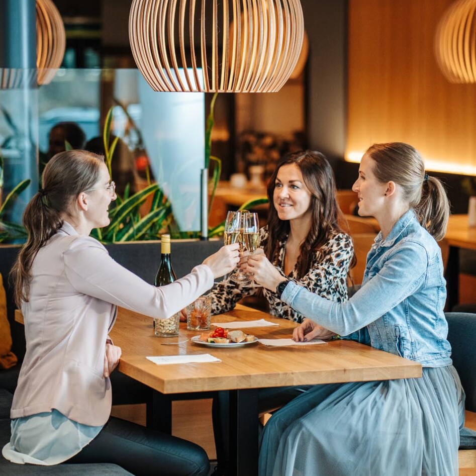 Drei Frauen stoßen mit Sektgläsern an. Eine elegante Lampe hängt über dem Tisch.