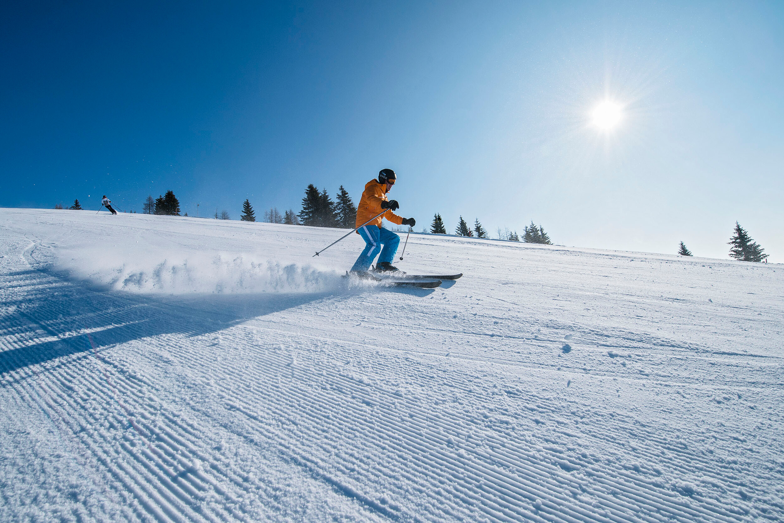 Ein Skifahrer in bunter Kleidung fährt bei Sonnenschein eine schneebedeckte Piste hinab, umgeben von Bäumen und blauem Himmel.